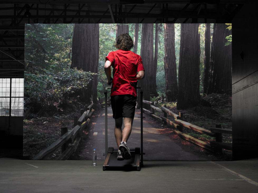 Young man on treadmill jogging in front photograph of forest