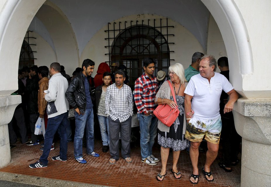 A tourist couple (R) and a group of temporarily detained immigrants take shelter from rain outside a police station in Kos island, Greece, in this May 29, 2015 file photo.  Italy stepped up calls for a change to European asylum rules on Sunday as neighbouring states tightened border controls, turning back African migrants and leaving hundreds stranded at the frontier in northern Italy.  REUTERS/Yannis Behrakis/Files ATTENTION EDITORS - THIS PICTURE IS PART OF THE PACKAGE "IN PLAIN SIGHT". TO FIND ALL 9 IMAGES SEARCH 'MIGRANT EUROPEAN'.