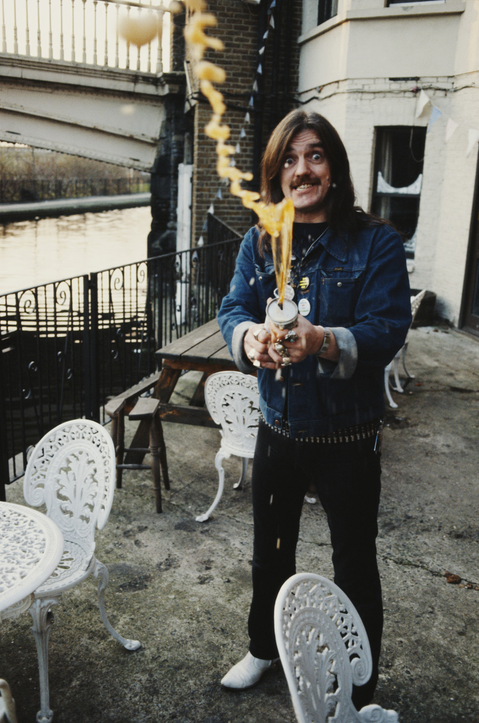 English rock musician and lead vocalist of the rock and roll band Motörhead, Lemmy (Ian Fraser 'Lemmy' Kilmister), spraying beer outside the Carlton Bridge Tavern, Grand Union Canal, London, circa 1985. (Photo by Tony Mottram/Getty Images)