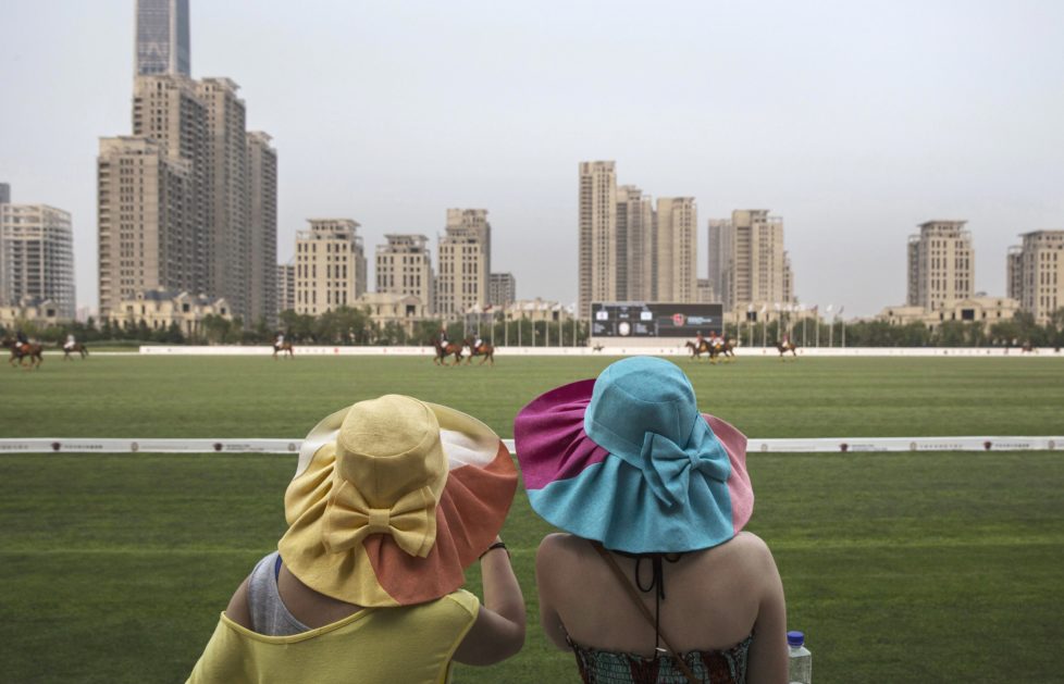 TIANJIN, CHINA - JULY 17: Chinese spectators watch a match between Chinese players from the Metropolitan Polo Club team and visiting players from the United States and Britain during the intervarsity tounrament at the Tianjin Goldin Metropolitan Polo Club on July 17, 2016 in Tianjin, China. China's rising affluence has nurtured growing interest in polo and other past-times regarded as noble or prestigious by the country's elite. Clubs and international-size polo fields have been built in various cities including Beijing and Shanghai, and on the outskirts of Tianjin, where membership at the exclusive Goldin Metropolitan, China's largest polo club, is by invitation-only and fees can be significant for polo team owners. Increasingly, wealthy Chinese parents are choosing polo and other equestrian activities for their children as a way to bolster their credentials for admission to top-tier universities in the United States and the United Kingdom. While the so-called "sport of kings" became a mainstay in Hong Kong during the era of British rule, polo is a relatively new sport to mainland China. Professional polo players are frequently flown in from countries such as New Zealand and Argentina in order to field competitive matches. Many of the polo clubs in China are tied to luxury real estate developments. (Photo by Kevin Frayer/Getty Images)