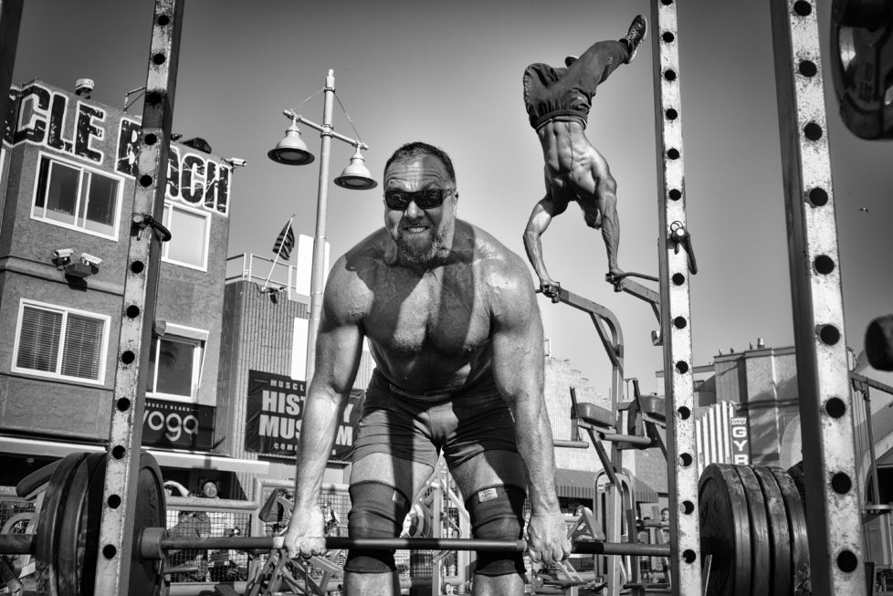 A weightlifter lifts a barbell loaded with heavy plates while a bodybuilder performs an aerial handstand at the Muscle Beach Gym in Venice Beach, CA.