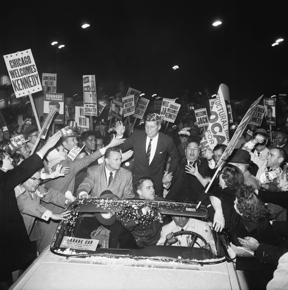 Democratic presidential candidate John F. Kennedy campaigns with Chicago mayor Richard J. Daley as a crowd of supporters swarms around his motorcade.