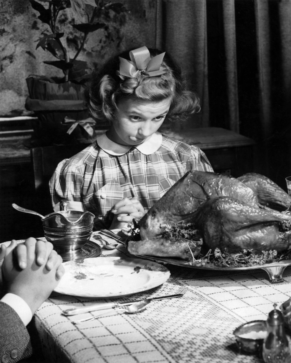 A young girl in a gingham dress with a white collar and a hair ribbon eyes the Thanksgiving turkey hungrily whilst prayers are being said around the table prior to Thanksgiving dinner in the USA, circa 1950. (Photo by Popperfoto/Getty Images)