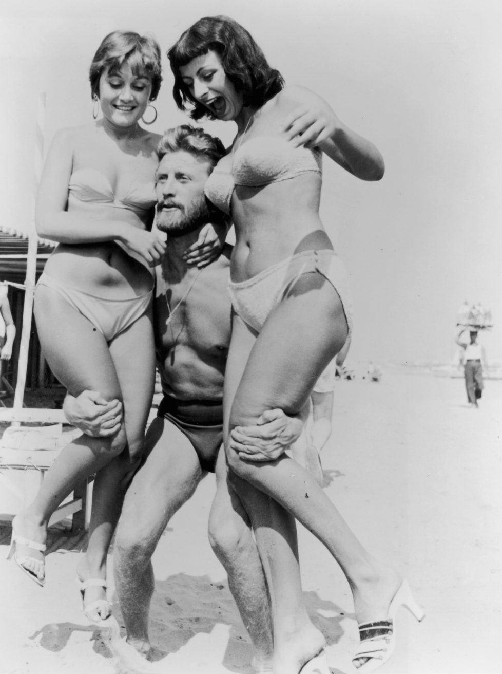 1953: American actor Kirk Douglas, wearing a swimsuit, lifts two women wearing bikinis on a beach during the Venice Film Festival, Italy. (Photo by Hulton Archive/Getty Images)