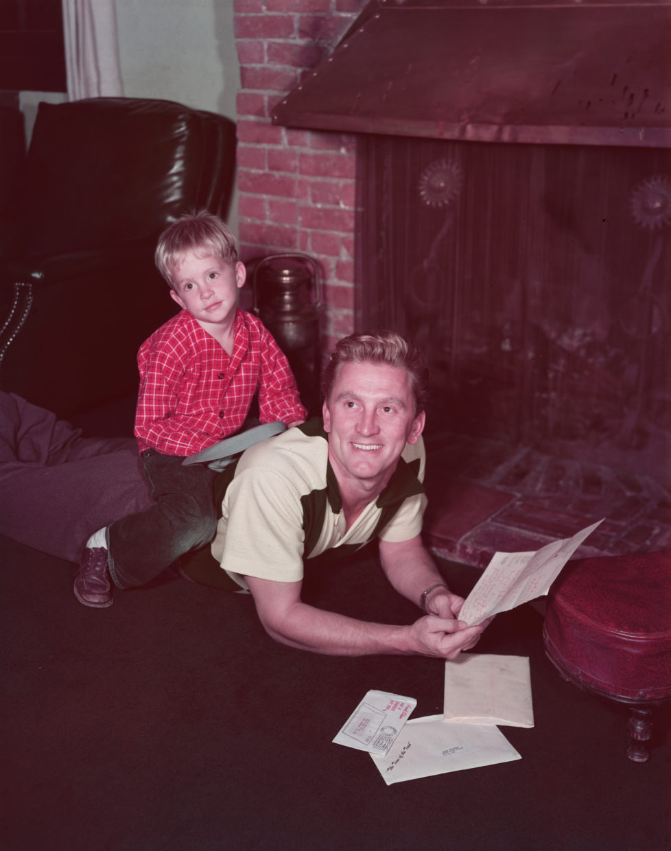 circa 1955: American actor Michael Douglas as a young boy, lounging with his father, the American actor Kirk Douglas, in front of a brick fireplace. Kirk is lying on his stomach and reading his mail as Michael sits on his back. (Photo by Hulton Archive/Getty Images)